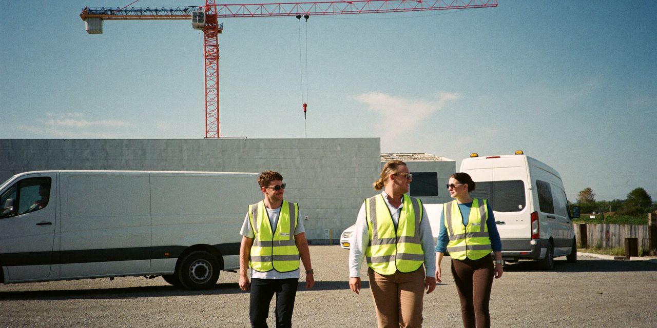 Technicians in hi-vis vests walking across a gravel yard from white vans at a data centre building site near Milan