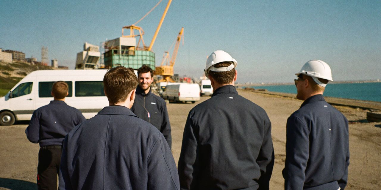 Electricians in navy work jackets talking at a harbour construction site with cranes and white vans